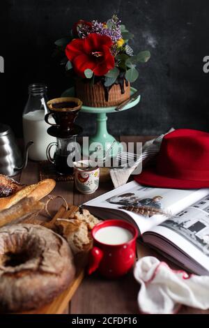 Zusammensetzung mit schönen Schokoladenkuchen mit frischen Blumen geschmückt serviert In der Nähe frisches Brot und Kaffee mit Milch auf Holztisch Mit Red hat und Buch Stockfoto