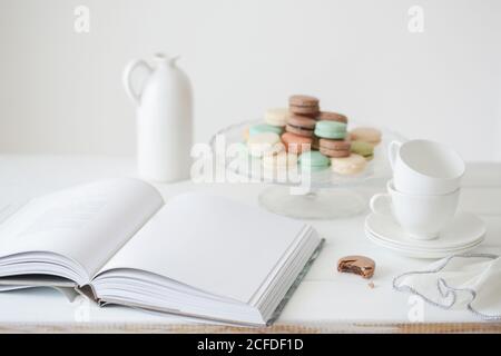 Von oben geöffnetes Buch mit leerer weißer Seite platziert Auf dem Tisch mit Teller mit bunten Makronen und Tee-Set Gegen weiße Wand Stockfoto