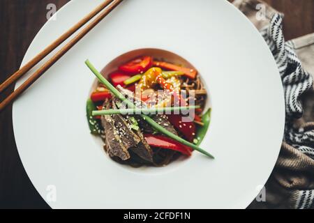 Draufsicht auf appetitlich würzige asiatische gebratene Nudeln mit Fleisch Und buntes Gemüse in weißem Teller mit Bambus-Essstäbchen serviert Auf Holztisch Stockfoto
