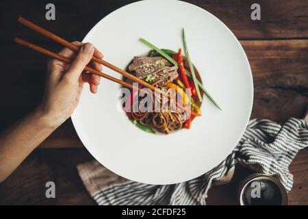 Draufsicht auf Erntegut Person mit Essstäbchen essen köstliche asiatische Nudeln mit geschnittenem Fleisch und buntem Gemüse in Weiß serviert Teller auf Holztisch Stockfoto