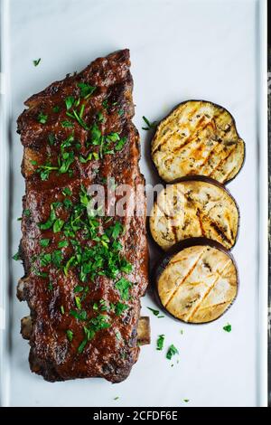 Blick von oben auf köstliche gegrillte Schweinerippchen garniert mit frischen Petersilie serviert mit gegrillter Aubergine in Scheiben auf weißem Tablett Stockfoto