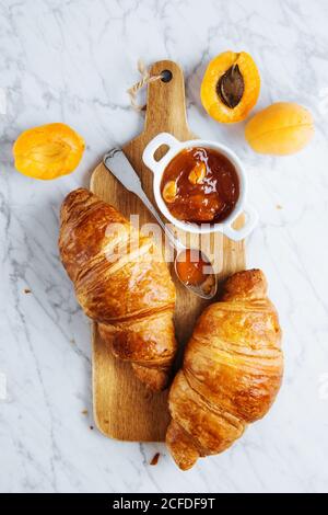 Blick von oben auf appetitlich frisches Croissant serviert mit Kanne Hausgemachte Aprikosenmarmelade auf Holz Schneidebrett in der Nähe frisch platziert Früchte auf Marmorhintergrund Stockfoto