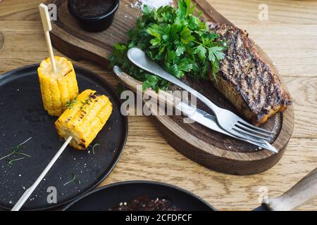Von oben köstliche gegrillte Steak mit frischem Grün serviert Kräuter und gegrillter Mais auf Stöcken auf Holztisch Stockfoto