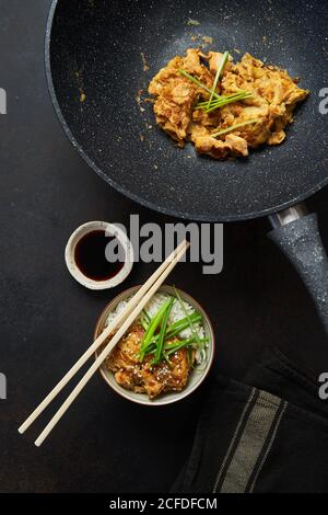 Draufsicht auf gebratenes Huhn auf Pfanne und Schüssel mit Japanische traditionelle Gericht Oyakodon serviert mit gekochtem Reis frische Kräuter sojasauce und Essstäbchen Stockfoto