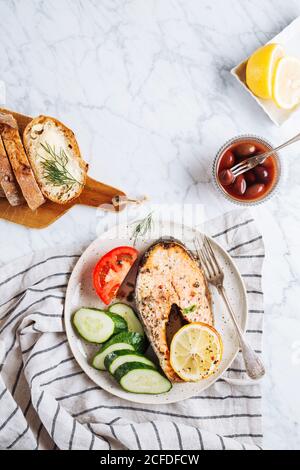 Blick von oben auf ein köstliches gesundes Abendessen mit aromatischem gegrilltem Lachs Steak mit Zitrone und Kräutern serviert mit frisch geschnittenen Gurken Und Tomaten auf Marmortisch mit frischem Brot und Oliven Stockfoto