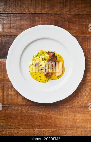 Draufsicht auf gegrilltes Fleisch auf weißem Teller, der gelb ausgießt Dicke Sauce mit Kräutern auf Holztisch im Restaurant Stockfoto
