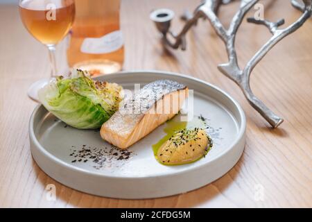 Gedämpfter Lachs mit gegrilltem Kohl auf weißem Keramikplatte serviert Auf Holztisch mit Glas und Flasche Wein Stockfoto