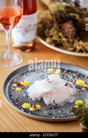 Stroganina oder Carpaccio mit frisch geschnittenem weißem Fisch serviert Auf silbernem Teller mit Soße und Salzflocken verziert Holztisch mit Rosenwein und Gericht im Hintergrund Stockfoto