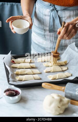 Von oben Ernte Koch hält weiße Schüssel und fleißig Bürsten Leckere Croissants auf Backblech auf dem Tisch in der Küche Stockfoto