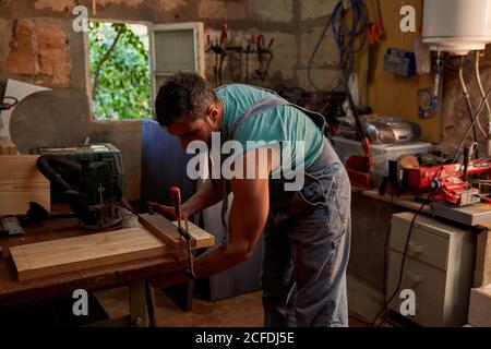 Seitenansicht des erfahrenen Handwerkers mittleren Alters in Overalls mit Klammer bei der Arbeit mit Holzbrett in der Werkstatt Stockfoto