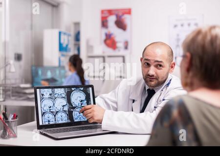 Arzt, der mit älteren Frauen mit alzheimer im Krankenhausbüro diskutiert und auf den Laptop-Bildschirm mit CT-Scan zeigt. Medic trägt weißen Mantel. Krankenschwester in blauer Uniform arbeiten am Computer. Stockfoto
