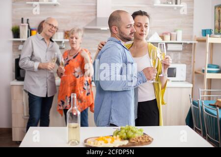 Ein Paar hält während des Brunchs mit der Familie in der Küche Weingläser und schaut sich diese an. Ältere Mutter und Vater schauen auf Sohn und seine Frau. Stockfoto