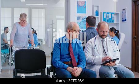 Arzt erklärt dem leitenden Mann im Wartebereich des Krankenhauses die Testergebnisse. Behinderte ältere Frau mit Gehrahmen Arring für die medizinische Untersuchung. Stockfoto