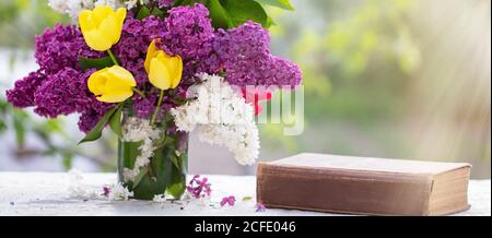 Federhintergrund. Flieder und Tulpen in einem Glas auf dem Tisch, auch ein altes Buch. Schöne Blumen, die von den Strahlen der Sonne beleuchtet werden. Stockfoto