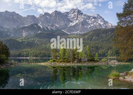 Blick über die Braxeninsel am Eibsee im Frühjahr Richtung Zugspitze, höchster Berg Deutschlands, türkisfarbenes Wasser, blauer Himmel, Wolken, Bäume, Stockfoto