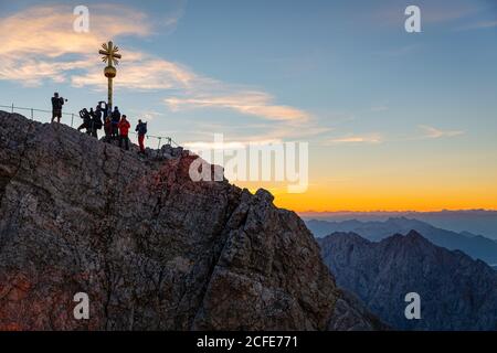 Mann mit Trompete spielt für Gruppe von Menschen auf Zugspitzgipfel vor Gipfelkreuz bei Sonnenaufgang, blauer Himmel, Wolken, Zugspitze, Grainau, Stockfoto