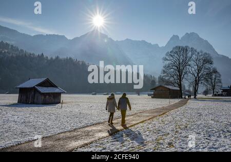 Mann und Frau bei einem Winterspaziergang auf einer Schotterstraße in Garmisch-Partenkirchen, mit Blick auf das Wettersteingebirge mit Alpspitze und Wachssteinen, Stockfoto