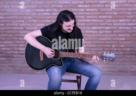 Junger männlicher Darsteller spielt Gitarre auf einem Stuhl sitzend. Stockfoto
