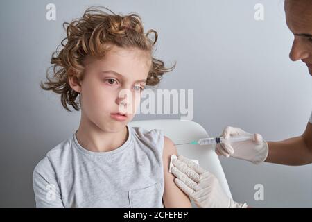 Crop Krankenschwester in weißen Latex-Handschuhe halten Schulter des Jungen Mit lockigen Haaren, während Impfstoffinjektion mit Spritze in Moderne Klinik Stockfoto