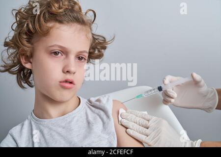 Crop Krankenschwester in weißen Latex-Handschuhe halten Schulter des Jungen Mit lockigen Haaren, während Impfstoffinjektion mit Spritze in Moderne Klinik Stockfoto