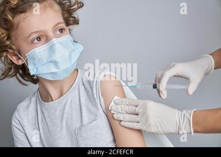 Crop Krankenschwester in weißen Latex-Handschuhe halten Schulter des Jungen Mit lockigen Haaren, während Impfstoffinjektion mit Spritze in Moderne Klinik Stockfoto