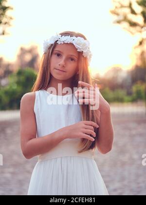 Kleine niedliche Mädchen in weißen eleganten Kleid und Blume Stirnband In der Mitte eines Parks stehend, während man anschaut Kamera Stockfoto