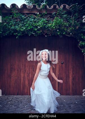 Nettes kleines Mädchen in weißen langen Kleid und Stirnband springen In der Nähe einer Holztür mit Efeu, der die Kamera anschaut Stockfoto