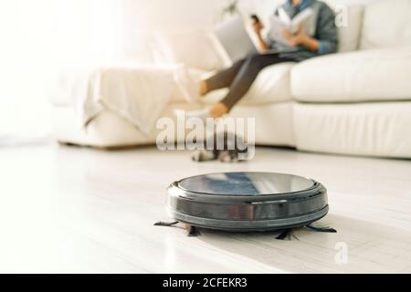 Niedlicher Welpe, der sich am nächsten Platz in einem schwarzen Roboter-Staubsauger befindet, während die Frau auf dem Sofa in einem hellen Raum mit Laminatboden sitzt Stockfoto