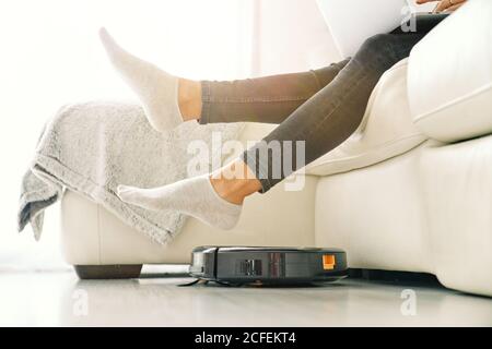 Seitenansicht der Crop-Frau in Jeans und Socken sitzen Auf weißem Sofa im Zimmer mit Laminatboden und Putting Füße am Roboter-Staubsauger Stockfoto