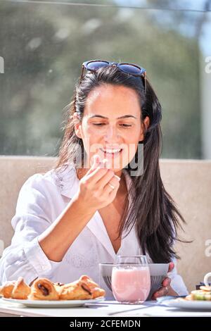 Ziemlich freundliche Dame, die am servierten Tisch auf offener Sonnenbeleuchtung saß Terrasse und essen rosa Joghurt, während auf Partner warten Stockfoto