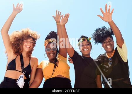 Niedriger Winkel der Gesellschaft von freundlichen multiethnischen Frauen, die Freude ausdrücken Mit erhobenen Armen auf dem Hintergrund des wolkenlosen Himmels im Sommer Und Blick auf die Kamera Stockfoto