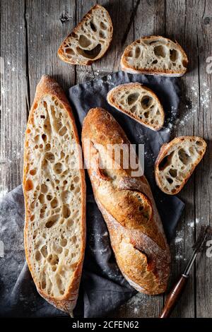 Von oben ganze und halbierte appetitliche Baguettes auf Weiß angeordnet Handtuch vor rustikalem Holzhintergrund Stockfoto