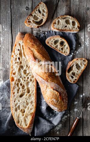 Von oben ganze und halbierte appetitliche Baguettes auf Weiß angeordnet Handtuch vor rustikalem Holzhintergrund Stockfoto