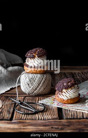 Hausgemachte Choux Gebäck mit Sahne und Schokolade Schere und Ball Von Garn auf Textur Holzoberfläche auf schwarzem Hintergrund angeordnet Stockfoto