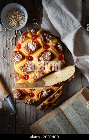 Blick von oben auf frisches Coca de San Juan Gebäck mit Samen und offenes Rezeptbuch auf Holztisch in der Nähe platziert Handtuch und Messer in rustikaler Küche Stockfoto