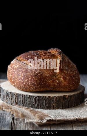 Laib frisches Land Sauerteig Brot auf Stück gelegt Holz auf schäbigen Tisch vor schwarzem Hintergrund Stockfoto