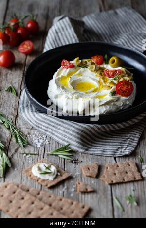 Knusprige Cracker und Rosmarin auf Holztisch neben Serviette gelegt Und Teller Labneh Joghurt mit Tomaten und Oliven Stockfoto