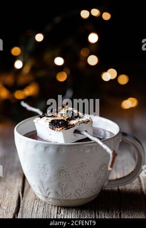 Geröstete Marshmallow auf Stick über elegante Tasse heiß platziert Schokolade auf Holztisch mit funkelnden dunklen Hintergrund Stockfoto