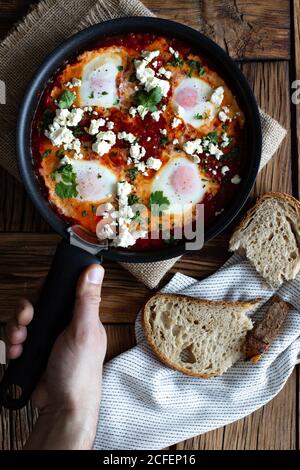 Draufsicht auf Erntegut Person auf Holztisch Braten setzen Pfanne mit leckeren Shakshuka mit Spiegeleiern mit Gemüse gemacht Und Feta-Käse Stockfoto