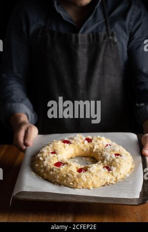 Crop Chef in schwarzer Schürze hält ungebackenes rundes Brot gekrönt Mit Kirsche beim Stehen am Holztisch Stockfoto