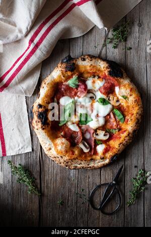 Blick von oben auf köstliche frische Sommerpizza auf Holz gelegt Tisch neben Handtuch und Schere in rustikaler Küche Stockfoto