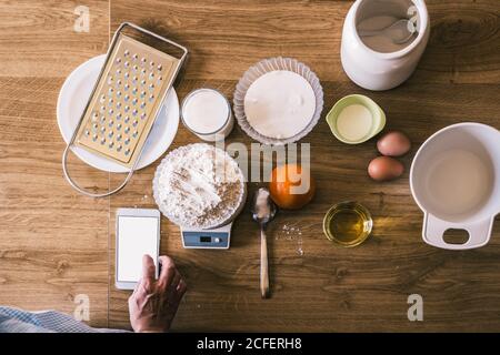 Draufsicht der Ernte anonyme weibliche Browsing Rezept auf dem Smartphone Und Weizenmehl auf elektronischen Waagen wiegen, während Zutaten zubereitet werden Für hausgemachtes Gebäck in der Küche Stockfoto