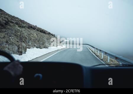 Hand der anonymen Person, die Auto auf Asphaltstraße durch Schneebedecktes bergiges Gelände an nebligen Tagen Stockfoto