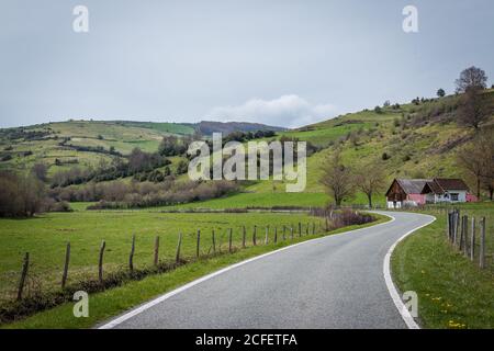 Enge kurvenreiche Straße in der Nähe von Bauernhäusern und grünen Rasen Hügel an bewölkten Tag in der Landschaft Stockfoto