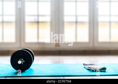 Von oben Sport-Set für das Training bestehend aus schwarz zusammenklappbar Kurzhantel und weißes Handtuch auf hellblauer Sportmatte auf Boden vor verschwommener Fensterwand im geräumigen modernen Fitnessraum Stockfoto