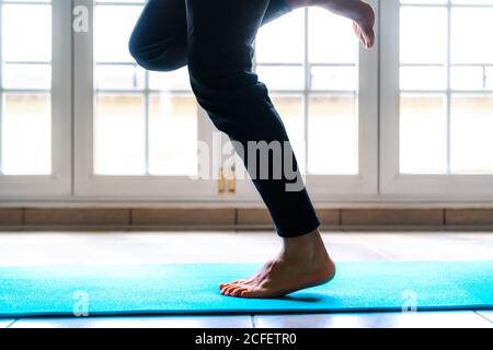 Low-Angle-Seitenansicht des Crop-fokussierten männlichen Athleten in Sport Kleidung tun einzelne Bein springen Übung auf lebendige blau Sportmatte im hellen, geräumigen Fitnessraum Stockfoto