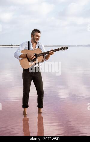 Leidenschaftlicher Mann in weißem Hemd und Hosenträger spielen Gitarre, während Barfuß im Wasser am Ufer am bewölkten Tag stehen Stockfoto