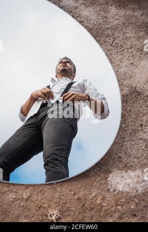 Low Angle Reflexion von verträumten Mann in Shirt und Hosenträger Über blauem Himmel in ovalem Spiegel auf staubigen Boden stehend Stockfoto