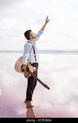 Melancholischer leidenschaftlicher Mann in weißem Hemd und Hosenträger mit Gitarre Während Sie barfuß im Wasser stehen und die Hand zum Himmel heben Auf dem Land Stockfoto