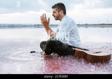 Mann in weißem Hemd und Hosenträger, der Wasser auf sein Gesicht wirft, während er am Strand mit rosa Wasser neben einer akustischen Gitarre sitzt, umgeben von einer sanften majestätischen Wolkenlandschaft Stockfoto